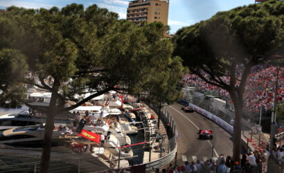 Race cars navigate a tight harbor street circuit as spectators fill grandstands; boats docked at left behind pine trees.