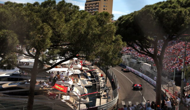 Race cars navigate a tight harbor street circuit as spectators fill grandstands; boats docked at left behind pine trees.