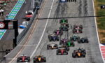 F1 cars line up and start a race on a gray circuit, tire marks visible on the track.