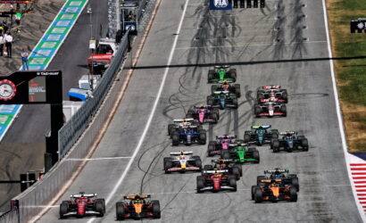 F1 cars line up and start a race on a gray circuit, tire marks visible on the track.