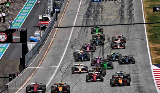 F1 cars line up and start a race on a gray circuit, tire marks visible on the track.