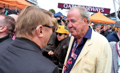 Two men in conversation at a crowded outdoor event with an orange tent and a Qatar Airways banner in the background.
