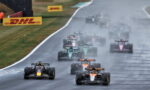 Formula 1 cars race on a rain-soaked track, spraying water as they speed toward the camera.