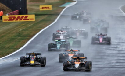 Formula 1 cars race on a rain-soaked track, spraying water as they speed toward the camera.