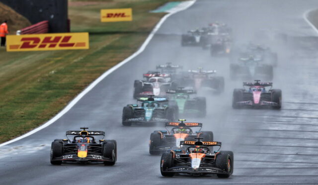 Formula 1 cars race on a rain-soaked track, spraying water as they speed toward the camera.