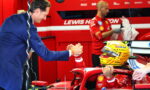 Ferrari Formula 1 driver in red suit sits in the cockpit of a red F1 car in the garage, helmet on, shaking hands with a team member in a blue suit.