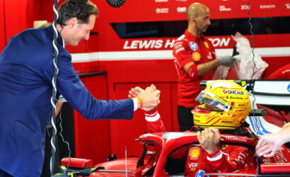 Ferrari Formula 1 driver in red suit sits in the cockpit of a red F1 car in the garage, helmet on, shaking hands with a team member in a blue suit.