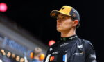 Male race car driver in a yellow and black cap and black sponsor-laden racing suit, looking off to the right at an indoor event.