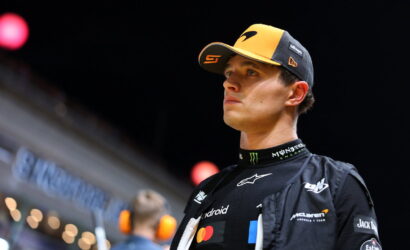 Male race car driver in a yellow and black cap and black sponsor-laden racing suit, looking off to the right at an indoor event.
