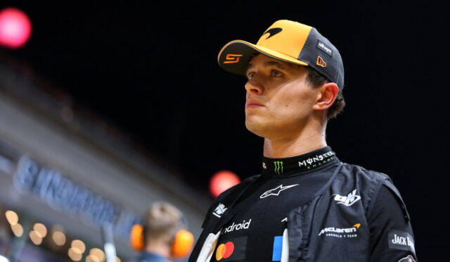 Male race car driver in a yellow and black cap and black sponsor-laden racing suit, looking off to the right at an indoor event.