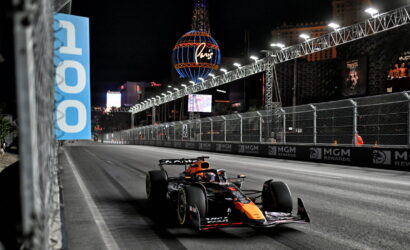 Nighttime Formula 1 car speeds along a barrier-lined Las Vegas street circuit, with bright lights and MGM Rewards banners along the track.