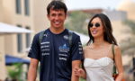 Smiling man in a dark blue racing jersey walks arm in arm with a woman in a white dress and sunglasses, outdoors.