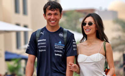 Smiling man in a dark blue racing jersey walks arm in arm with a woman in a white dress and sunglasses, outdoors.