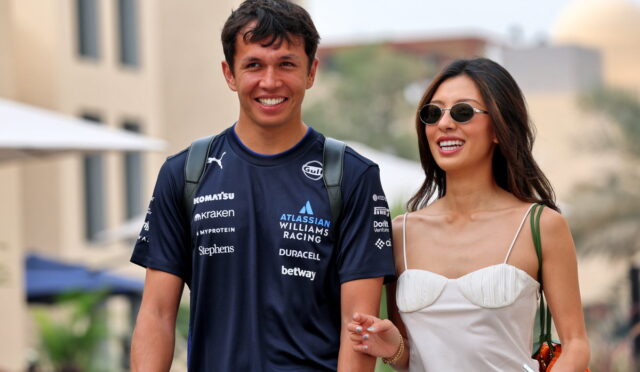 Smiling man in a dark blue racing jersey walks arm in arm with a woman in a white dress and sunglasses, outdoors.
