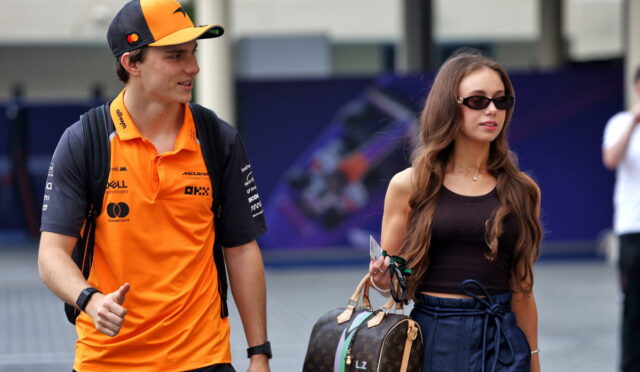 Young man in an orange polo and cap gives a thumbs up while walking beside a woman with sunglasses and a designer LV handbag outside.