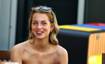 Smiling blonde woman with sunglasses on her head sits at a table outdoors, wearing a black strapless top.