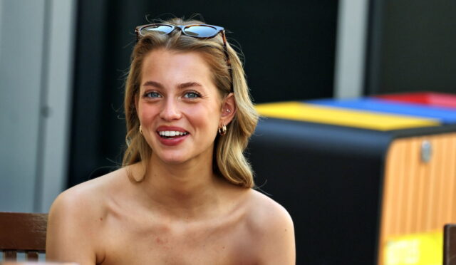 Smiling blonde woman with sunglasses on her head sits at a table outdoors, wearing a black strapless top.