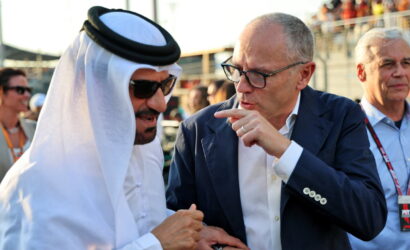 Two men converse at an outdoor event: one in a white traditional outfit and sunglasses, the other in a blue suit pointing as he speaks with him in the foreground.