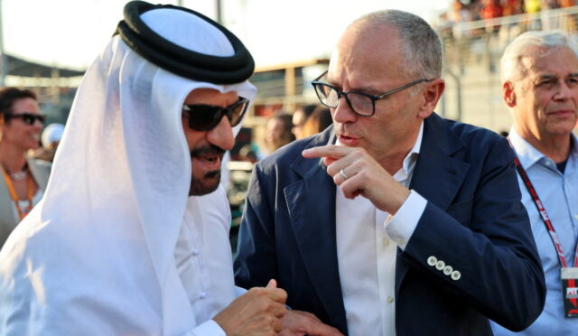 Two men converse at an outdoor event: one in a white traditional outfit and sunglasses, the other in a blue suit pointing as he speaks with him in the foreground.