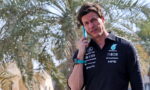 Man in a dark polo shirt with sponsor logos talks on a blue smartphone outdoors, palm trees behind him.