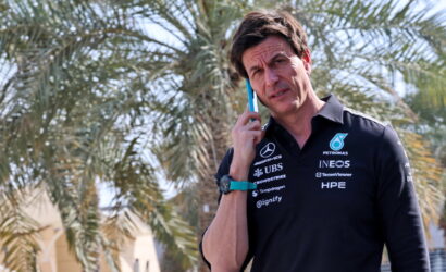 Man in a dark polo shirt with sponsor logos talks on a blue smartphone outdoors, palm trees behind him.