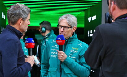Team member in a teal jacket being interviewed, holding a Sky Sport microphone, in a green-lit garage/pit area.
