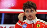 Formula 1 driver in a red Ferrari racing suit adjusts an earpiece against a red sponsor backdrop.