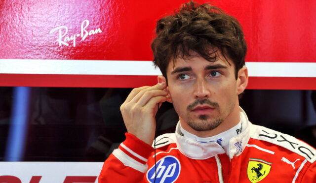Formula 1 driver in a red Ferrari racing suit adjusts an earpiece against a red sponsor backdrop.