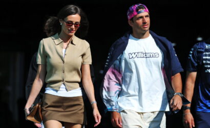 A stylish woman in a beige short-sleeve cardigan and brown mini skirt walks beside a man wearing a white Williams t-shirt and a pink cap, outdoors on a sunny day.