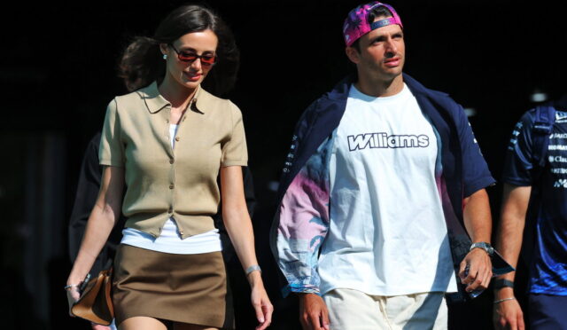 A stylish woman in a beige short-sleeve cardigan and brown mini skirt walks beside a man wearing a white Williams t-shirt and a pink cap, outdoors on a sunny day.