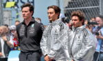 Three men stand side by side at a Formula 1 event; two wear silver jackets, one in a black team polo, with fans in the background.