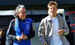Man in blue sponsor-laden racing suit smiles while walking with a casually dressed man in a light gray suit outside a pit area.