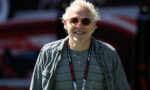 Smiling man with blond hair wearing sunglasses, a gray striped shirt, and a denim jacket at an outdoor event with a lanyard around his neck.