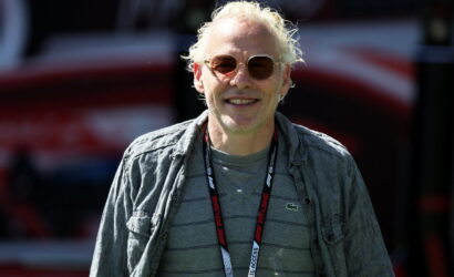 Smiling man with blond hair wearing sunglasses, a gray striped shirt, and a denim jacket at an outdoor event with a lanyard around his neck.