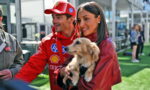 Man in red racing jacket and cap with sponsor logos stands beside a smiling woman holding a small beige dog at an outdoor event.