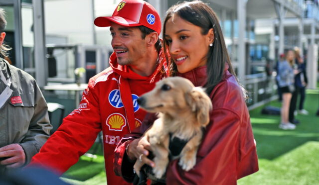 Man in red racing jacket and cap with sponsor logos stands beside a smiling woman holding a small beige dog at an outdoor event.
