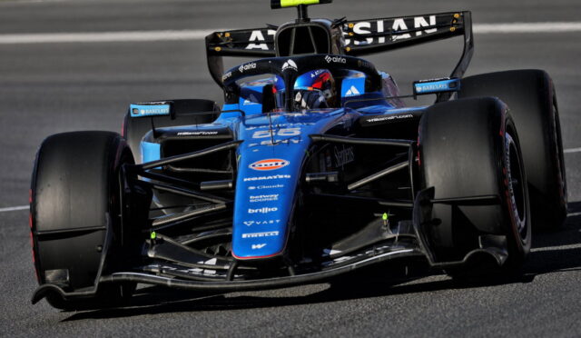 Blue open-wheel Formula 1 race car on a track, driver in helmet visible in the cockpit, front wing and aero complex in focus for a dynamic speed shot