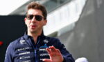 Male race car driver wearing dark sunglasses and a navy sponsor jacket, gesturing with his hand outdoors.