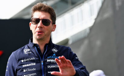 Male race car driver wearing dark sunglasses and a navy sponsor jacket, gesturing with his hand outdoors.