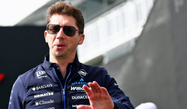 Male race car driver wearing dark sunglasses and a navy sponsor jacket, gesturing with his hand outdoors.
