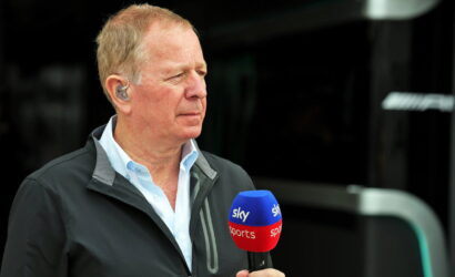 Side-profile of an older man in a light blue shirt and dark jacket, holding a blue and red Sky Sports microphone during an interview.