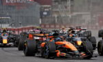 Orange Formula 1 car leads a pack on a track, with several rival cars close behind as barriers line the circuit.