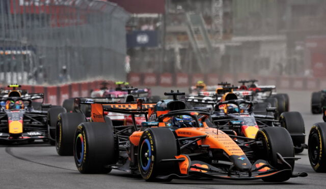 Orange Formula 1 car leads a pack on a track, with several rival cars close behind as barriers line the circuit.
