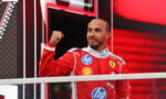 Ferrari race driver in red suit raises fist in victory gesture on a podium, smiling against a red, high-tech backdrop.