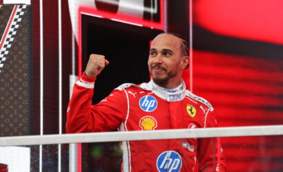 Ferrari race driver in red suit raises fist in victory gesture on a podium, smiling against a red, high-tech backdrop.