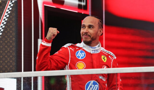 Ferrari race driver in red suit raises fist in victory gesture on a podium, smiling against a red, high-tech backdrop.
