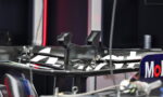 Close-up of a carbon fiber race car rear wing in a pit/garage, with sponsor decals on the endplate visible.