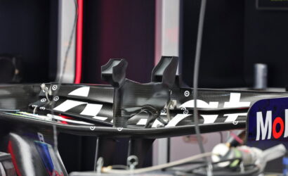 Close-up of a carbon fiber race car rear wing in a pit/garage, with sponsor decals on the endplate visible.