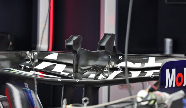 Close-up of a carbon fiber race car rear wing in a pit/garage, with sponsor decals on the endplate visible.