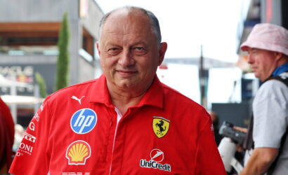 Smiling man in a red Ferrari team polo with sponsor logos (HP, Shell, UniCredit) outdoors in a paddock area.
